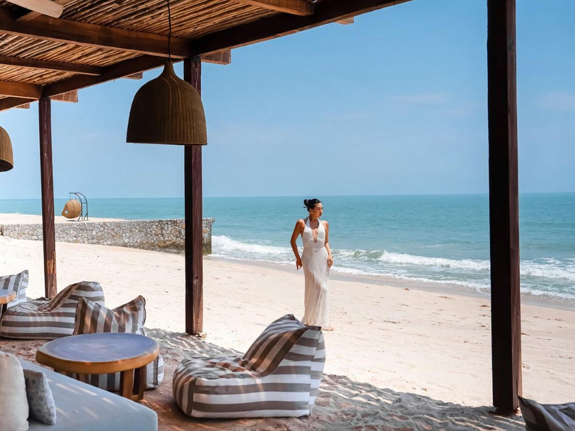 A woman in an elegant white halter-neck dress stands gracefully on a sandy beach by the turquoise ocean, with a striped beanbag chair and wooden pillars in the foreground.