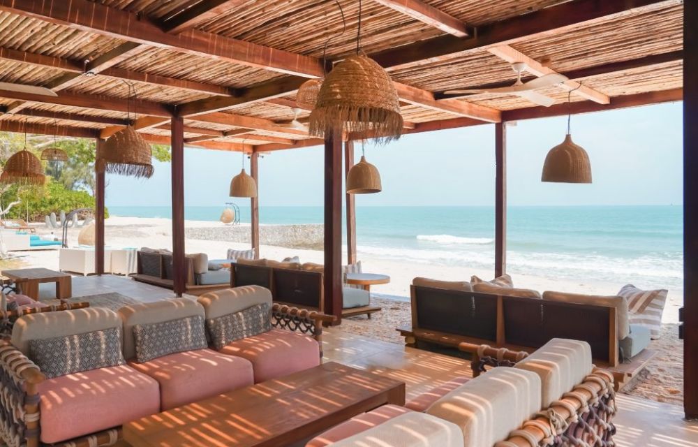 http://A%20rustic%20yet%20chic%20beachfront%20lounge%20restaurant%20with%20woven%20light%20fixtures,%20comfortable%20sofas%20and%20seating%20areas,%20offering%20open%20views%20of%20the%20ocean%20and%20sandy%20beach.