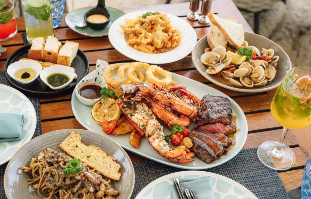 http://An%20elaborate%20gourmet%20platter%20featuring%20grilled%20lobster,%20king%20prawns,%20sliced%20steak,%20calamari,%20onion%20rings,%20and%20pasta,%20served%20on%20a%20wooden%20table%20with%20cocktails.