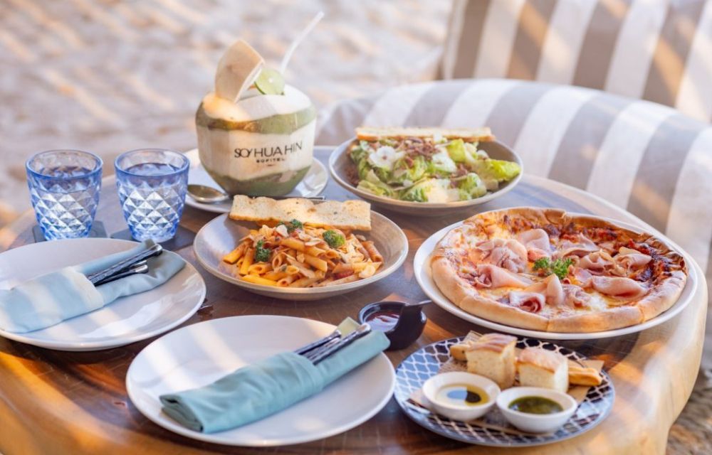http://A%20casual%20beachside%20dining%20setup%20on%20a%20low%20wooden%20table%20with%20a%20fresh%20pizza,%20Caesar%20salad,%20pasta,%20bread,%20and%20a%20coconut%20drink,%20perfect%20for%20a%20relaxed%20meal.