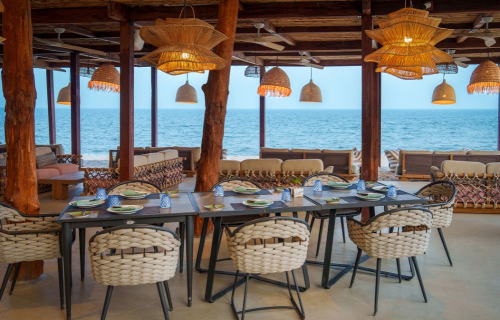 http://An%20elegant%20beachfront%20restaurant%20dining%20area%20with%20a%20long%20table%20set%20with%20patterned%20plates,%20woven%20chairs,%20and%20stylish%20pendant%20lights,%20offering%20an%20uninterrupted%20view%20of%20the%20ocean.
