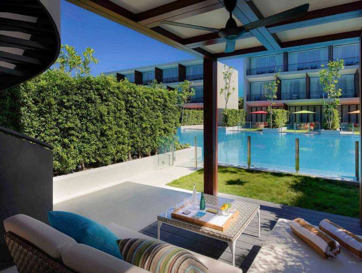 A spacious outdoor terrace with a comfortable daybed lounge area, a small table with refreshments, and a view of a private swimming pool, surrounded by lush green hedges.