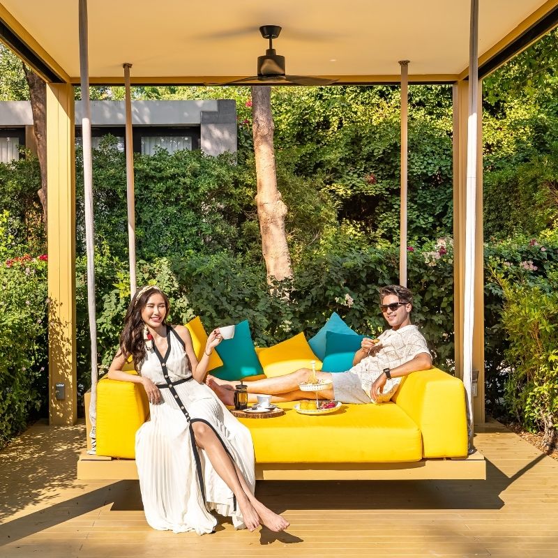A smiling couple relaxes on a bright yellow swinging daybed in an outdoor cabana, enjoying breakfast and coffee amidst lush tropical greenery on a sunny day.