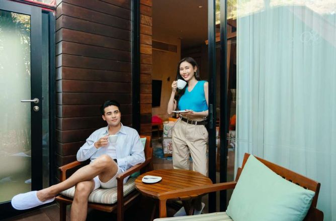http://A%20smiling%20couple%20sitting%20on%20a%20cozy%20hotel%20patio%20with%20wooden%20furniture,%20drinking%20coffee;%20the%20man%20in%20a%20chair,%20the%20woman%20leaning%20against%20the%20doorframe.