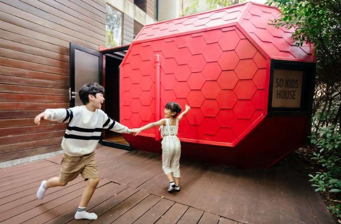 http://Two%20happy%20children%20holding%20hands%20and%20running%20outside%20a%20unique%20red,%20hexagonal%20playhouse%20labeled%20SO%20KIDS%20HOUSE%20on%20a%20wooden%20deck.