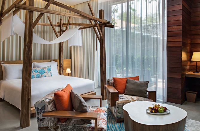 http://A%20luxurious%20hotel%20bedroom%20featuring%20a%20wooden%20canopy%20bed%20with%20white%20drapes,%20two%20colorful%20patterned%20armchairs%20with%20orange%20and%20gray%20cushions,%20and%20a%20white%20coffee%20table%20with%20fruit.