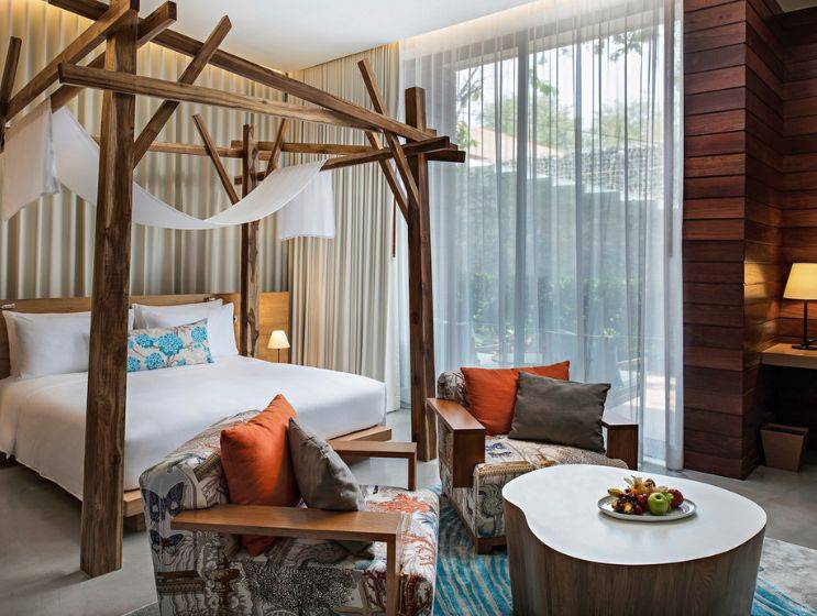 A luxurious hotel bedroom featuring a wooden canopy bed with white drapes, two colorful patterned armchairs with orange and gray cushions, and a white coffee table with fruit.