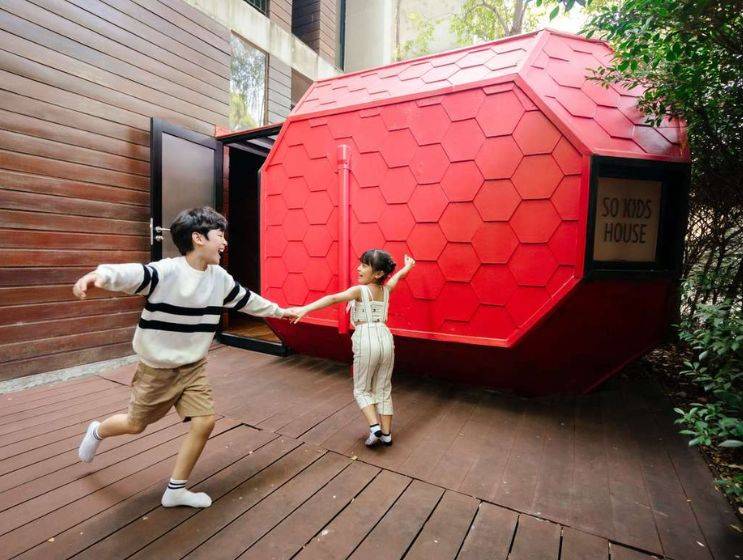 Two happy children holding hands and running outside a unique red, hexagonal playhouse labeled "SO KIDS HOUSE" on a wooden deck.