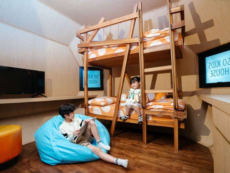 A fun, wood-paneled kids' room featuring a wooden bunk bed with orange polka-dot bedding, a blue beanbag chair where a boy reads, and a girl sitting on the top bunk.