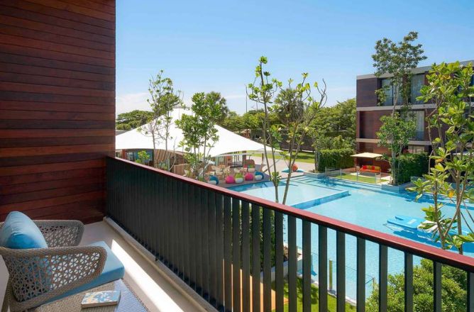 http://A%20hotel%20balcony%20with%20a%20wicker%20chair%20offering%20a%20panoramic%20view%20of%20a%20large%20resort%20swimming%20pool,%20a%20shaded%20play%20area%20with%20beanbags,%20and%20surrounding%20hotel%20buildings.