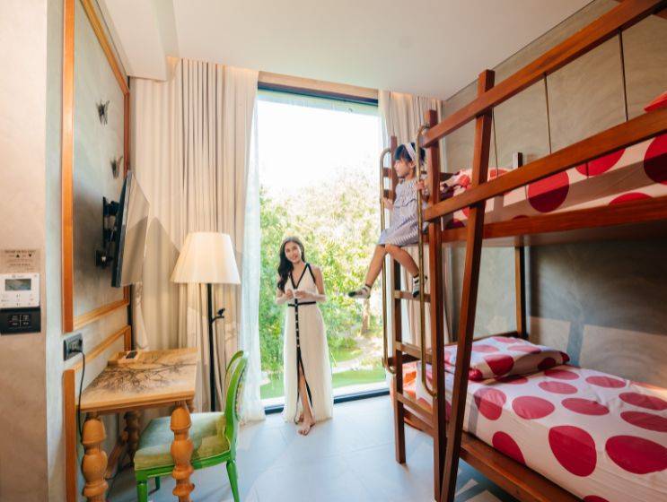A mother in a white dress stands by a large window in a family hotel room, looking at her daughter who is climbing into a wooden bunk bed with red polka-dot bedding.
