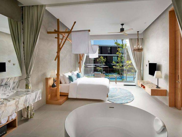 A luxurious hotel suite combining bedroom and bathroom, featuring a wooden canopy bed, a freestanding circular bathtub, a marble vanity, and a balcony overlooking a resort pool.
