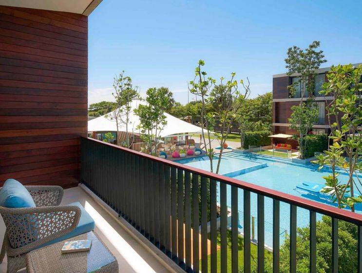 A hotel balcony with a wicker chair offering a panoramic view of a large resort swimming pool, a shaded play area with beanbags, and surrounding hotel buildings.