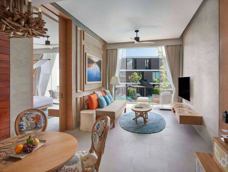 Bright and airy modern room featuring a cozy seating area, decorative art, and an inviting bedroom beyond, accented with vibrant pillows.