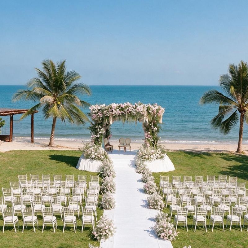 Beach wedding setup with a floral arch of pink and white flowers between two palm trees, surrounded by white chairs on green grass, overlooking the ocean.