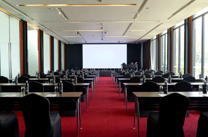 http://Conference%20room%20with%20rows%20of%20tables%20covered%20in%20black%20cloth%20and%20chairs%20facing%20a%20screen%20at%20the%20front.%20Large%20windows%20on%20the%20right%20let%20in%20natural%20light.