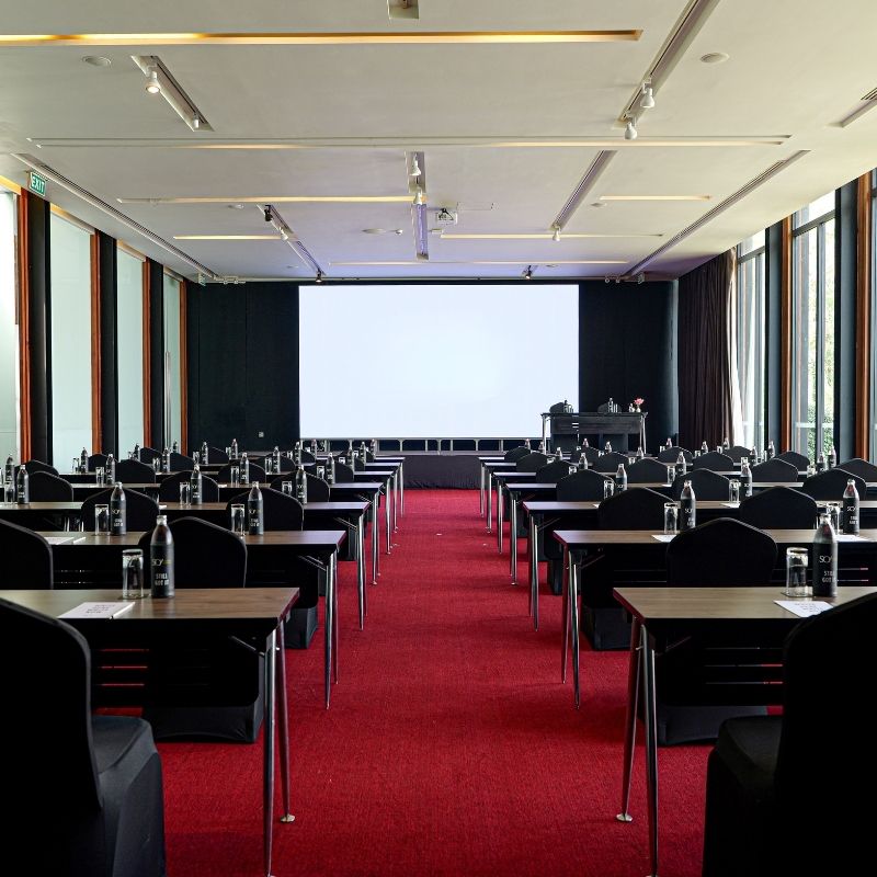 Spacious conference room with rows of tables and chairs, set with water bottles and notepads. Large windows let in natural light. A screen displays "SOFITEL AMBASSADOR."