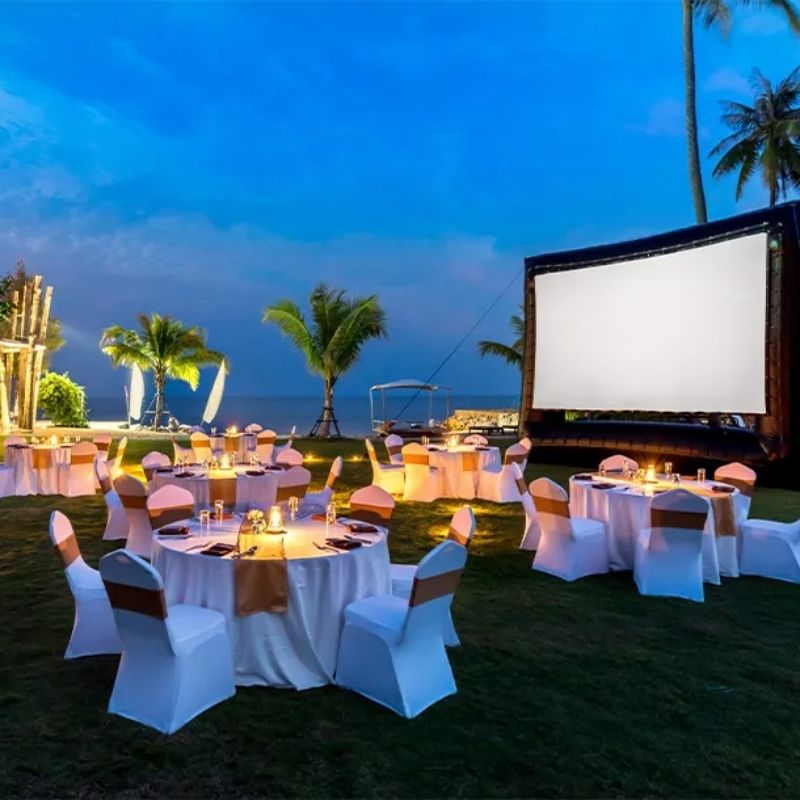 Outdoor evening event near a beach with round tables, white tablecloths, and candles. A large screen displays "SO HUA HIN" under palm trees.