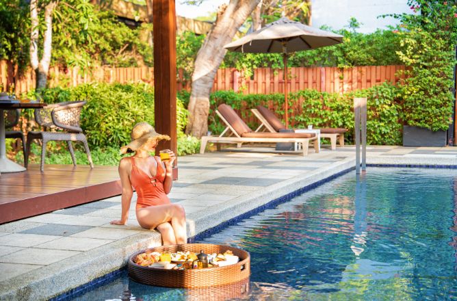 http://A%20woman%20in%20a%20swimsuit%20sits%20at%20the%20edge%20of%20a%20swimming%20pool,%20enjoying%20a%20floating%20breakfast%20tray%20with%20orange%20juice%20and%20food%20items,%20reflecting%20warm%20morning%20sunlight%20from%20a%20shaded%20wooden%20deck.