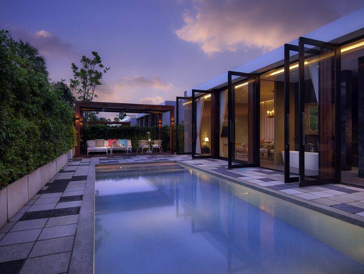 An inviting luxury private pool villa at dusk, with warmly lit interior rooms opening onto a tranquil swimming pool and a paved patio area, surrounded by lush trees.