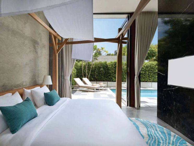A bright hotel bedroom with white bedding and a wooden canopy bed frame, featuring large glass doors that open directly onto a private pool area with outdoor seating.