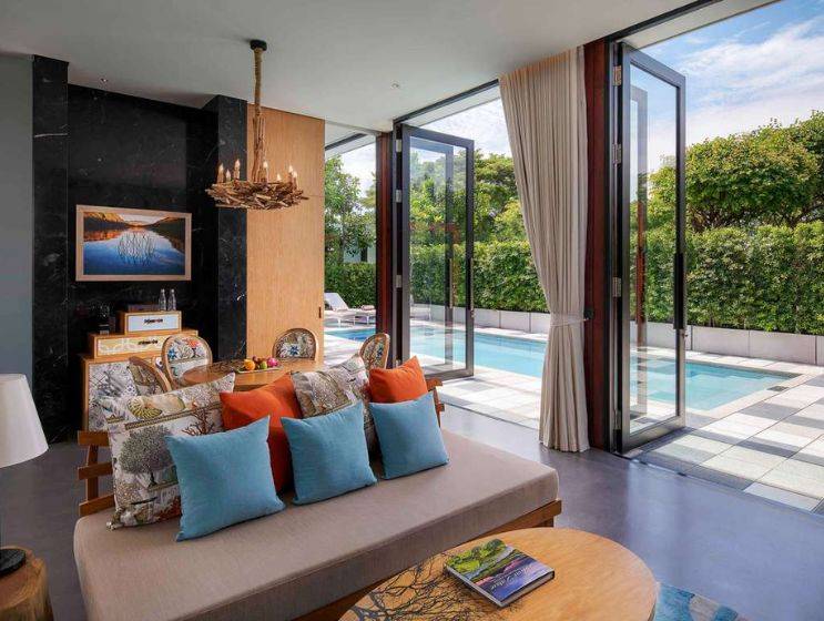 An elegant suite living area with a comfortable daybed, decorative cushions, and large folding glass doors that open wide to reveal a private swimming pool surrounded by greenery.