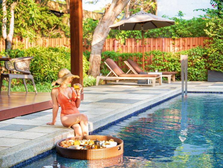 A woman in a swimsuit sits at the edge of a swimming pool, enjoying a floating breakfast tray with orange juice and food items, reflecting warm morning sunlight from a shaded wooden deck.