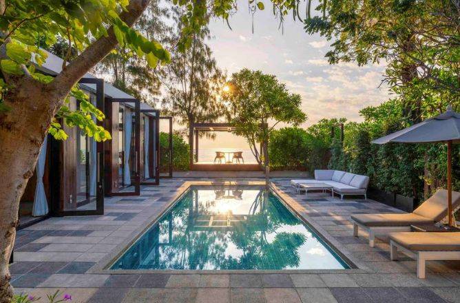 A serene outdoor scene featuring a turquoise pool beside a modern house with glass doors. A cozy patio area and lush greenery enhance the tranquil vibe.