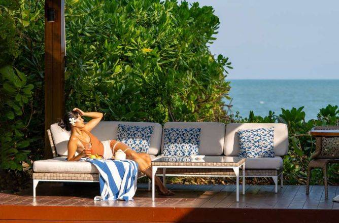 A woman in a swimsuit with a flower in her hair relaxes on a comfortable outdoor sofa on a wooden terrace, enjoying a refreshing drink with the ocean visible in the background.