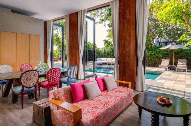A colorful hotel living room featuring a pink floral sofa, eclectic dining chairs, and large glass doors that open directly onto a private swimming pool and patio area.