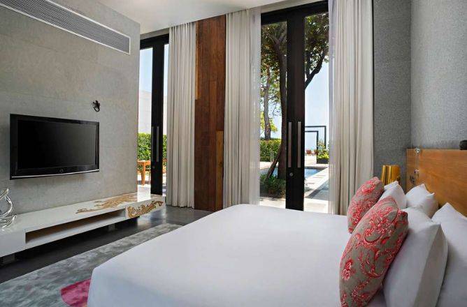 A sleek hotel bedroom with a large white bed, modern entertainment unit, and floor-to-ceiling glass doors providing direct views of a private pool and lush surroundings.