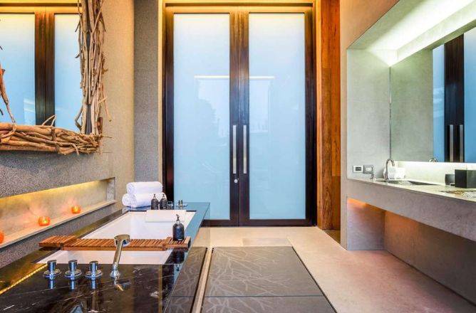 A luxurious spa-like bathroom featuring a deep soaking tub with a dark marble surround, a modern vanity with two sinks, and frosted glass doors leading outside.