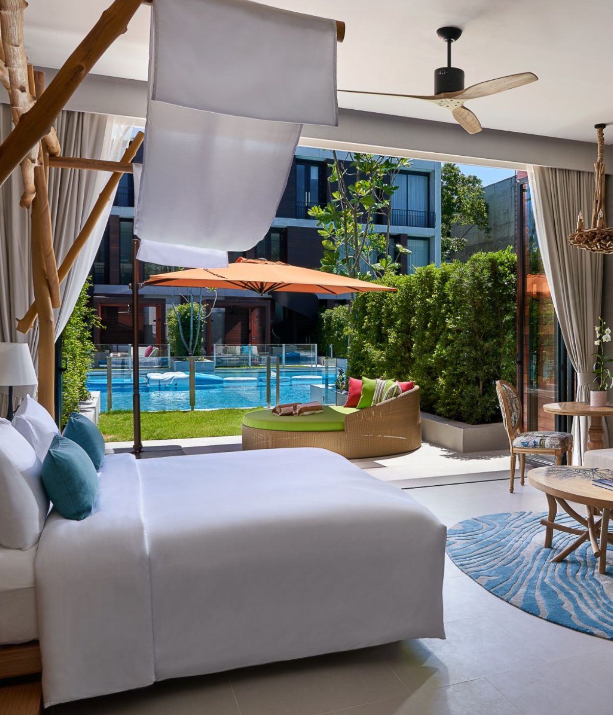 A bright hotel bedroom with white bedding and a wooden canopy bed frame, featuring large glass doors that open directly onto a private pool area with outdoor seating.