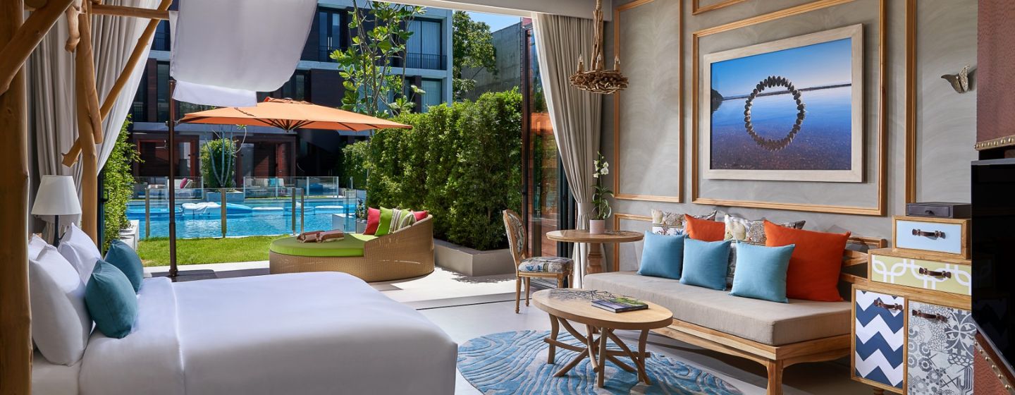 Modern hotel room with large bed, turquoise pillows, wooden accents, and blue rug. Open balcony shows pool view and greenery, creating a relaxing vibe.