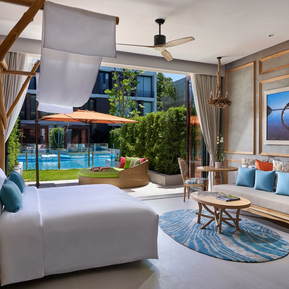 Modern hotel room with large bed, turquoise pillows, wooden accents, and blue rug. Open balcony shows pool view and greenery, creating a relaxing vibe.