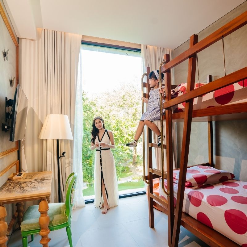 A mother in a white dress stands by a large window in a family hotel room, looking at her daughter who is climbing into a wooden bunk bed with red polka-dot bedding.