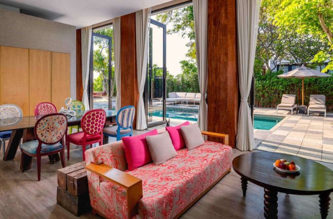 http://A%20colorful%20hotel%20living%20room%20featuring%20a%20pink%20floral%20sofa,%20eclectic%20dining%20chairs,%20and%20large%20glass%20doors%20that%20open%20directly%20onto%20a%20private%20swimming%20pool%20and%20patio%20area.
