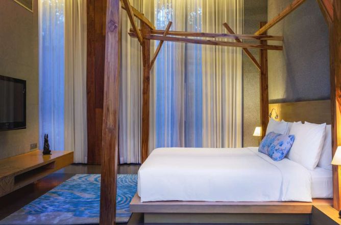 http://A%20modern%20hotel%20bedroom%20with%20a%20minimalist%20wooden%20bed%20frame,%20white%20bedding,%20and%20a%20light%20blue%20rug,%20featuring%20a%20large%20screen%20TV%20and%20tall%20sheer%20curtains%20behind%20the%20bed.