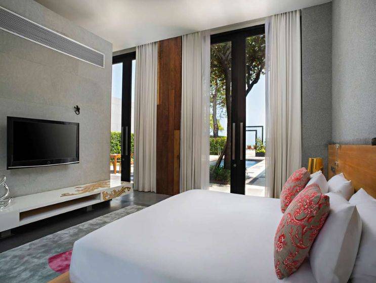 A sleek hotel bedroom with a large white bed, modern entertainment unit, and floor-to-ceiling glass doors providing direct views of a private pool and lush surroundings.