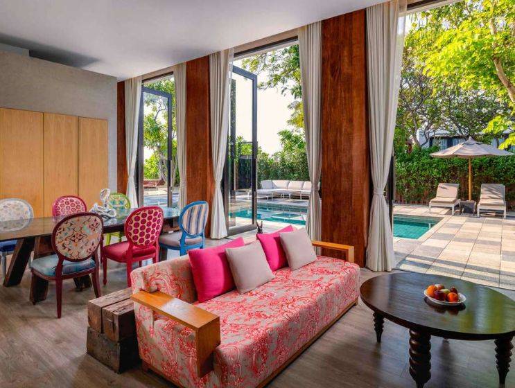 A colorful hotel living room featuring a pink floral sofa, eclectic dining chairs, and large glass doors that open directly onto a private swimming pool and patio area.