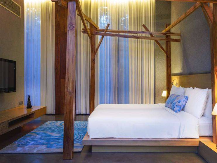 A modern hotel bedroom with a minimalist wooden bed frame, white bedding, and a light blue rug, featuring a large screen TV and tall sheer curtains behind the bed.