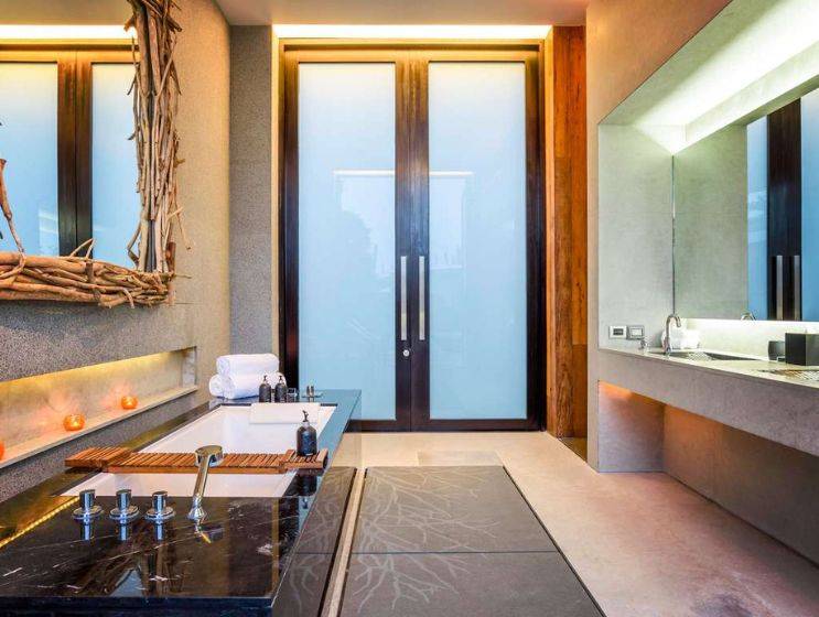 A luxurious spa-like bathroom featuring a deep soaking tub with a dark marble surround, a modern vanity with two sinks, and frosted glass doors leading outside.