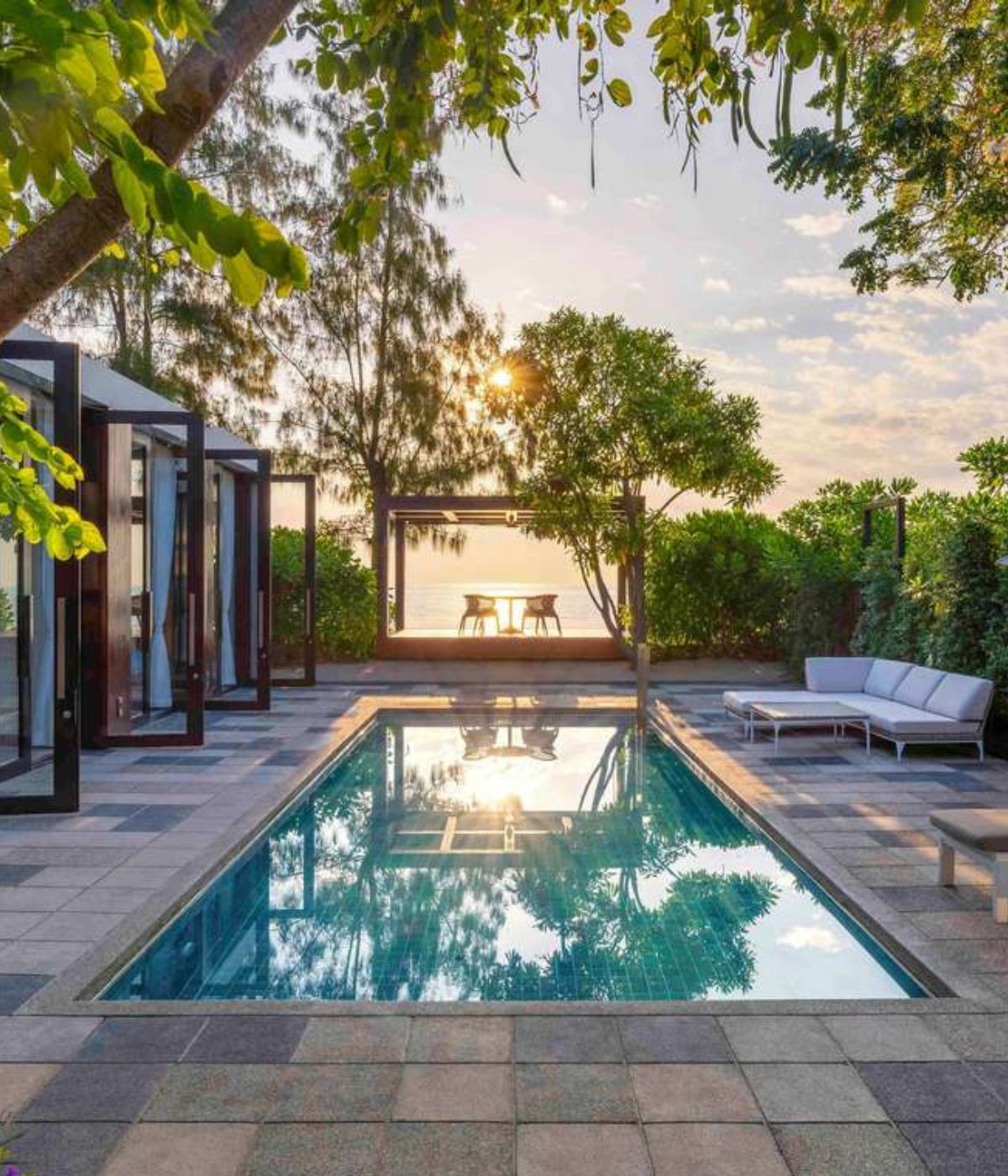 A serene outdoor scene featuring a turquoise pool beside a modern house with glass doors. A cozy patio area and lush greenery enhance the tranquil vibe.