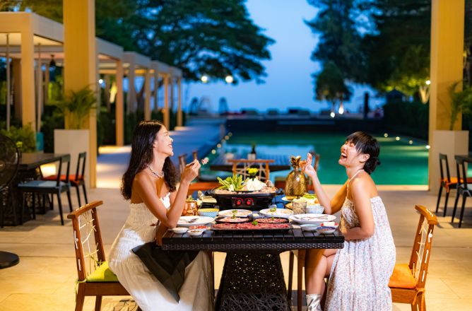 http://Two%20laughing%20women%20enjoying%20a%20hot%20pot%20dinner%20at%20a%20beautifully%20set%20outdoor%20table%20by%20the%20illuminated%20swimming%20pool%20at%20dusk,%20with%20a%20resort%20ambiance.