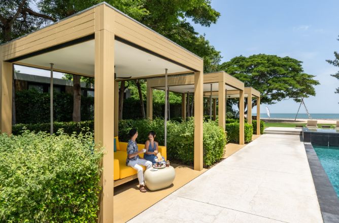 http://A%20couple%20enjoys%20breakfast%20or%20lunch%20in%20stylish%20yellow%20and%20beige%20resort%20cabanas%20by%20a%20swimming%20pool,%20surrounded%20by%20lush%20hedges%20under%20a%20clear%20blue%20sky.