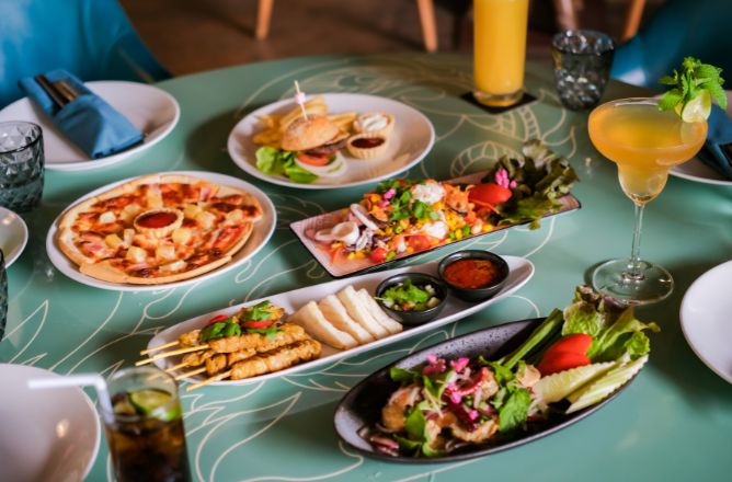 http://A%20vibrant%20dining%20table%20spread%20with%20diverse%20dishes%20including%20pizza,%20chicken%20skewers,%20a%20seafood%20salad,%20a%20mini-burger,%20and%20colorful%20cocktails%20on%20a%20light%20green%20patterned%20table.