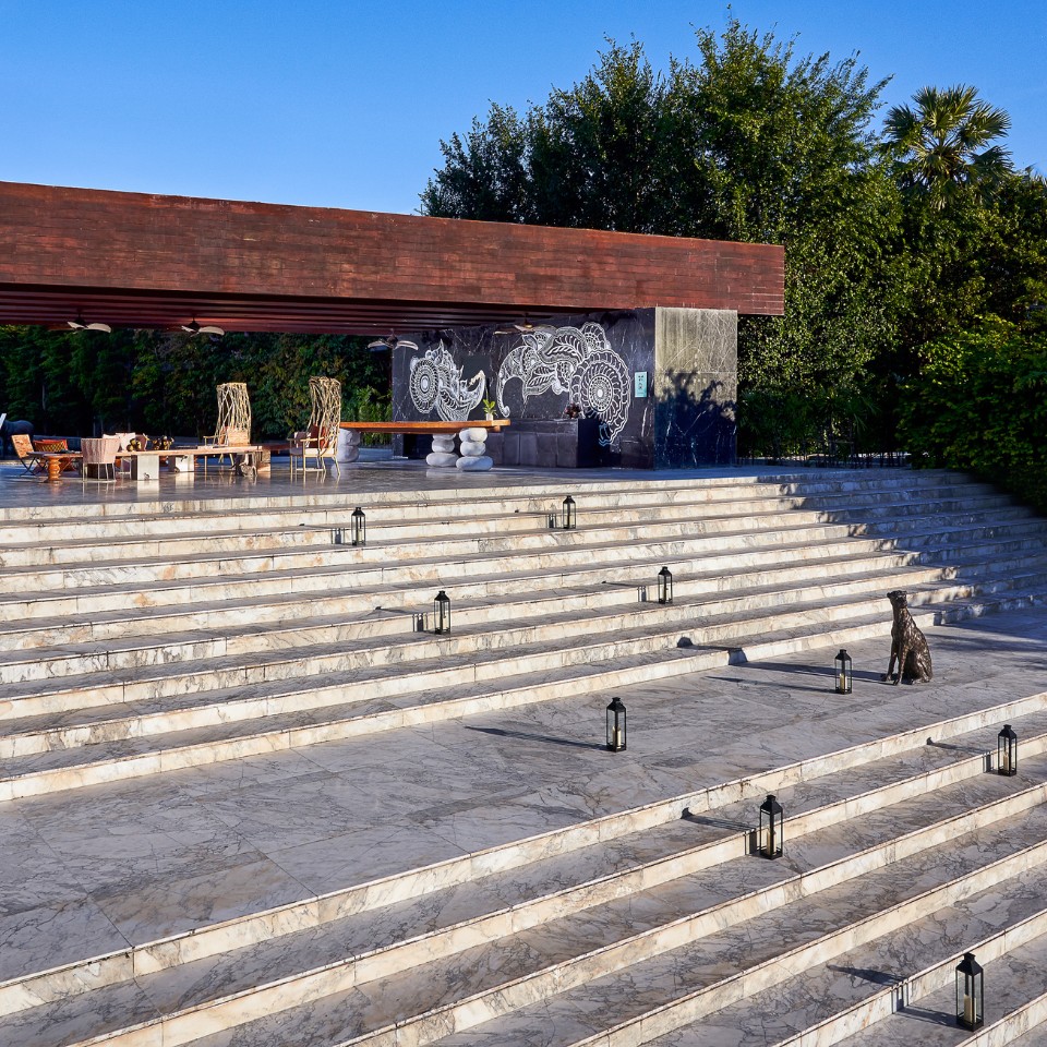 Wide marble steps lead to an outdoor pavilion with a wooden roof. The pavilion features abstract wall art and modern furnishings, set against lush greenery.