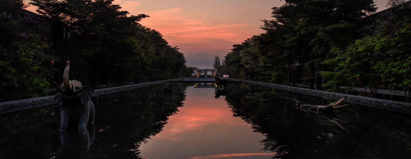 A tranquil pool reflects a vibrant pink and orange sunset, framed by dark trees. Elephant statues emerge from the water, adding a serene tone.