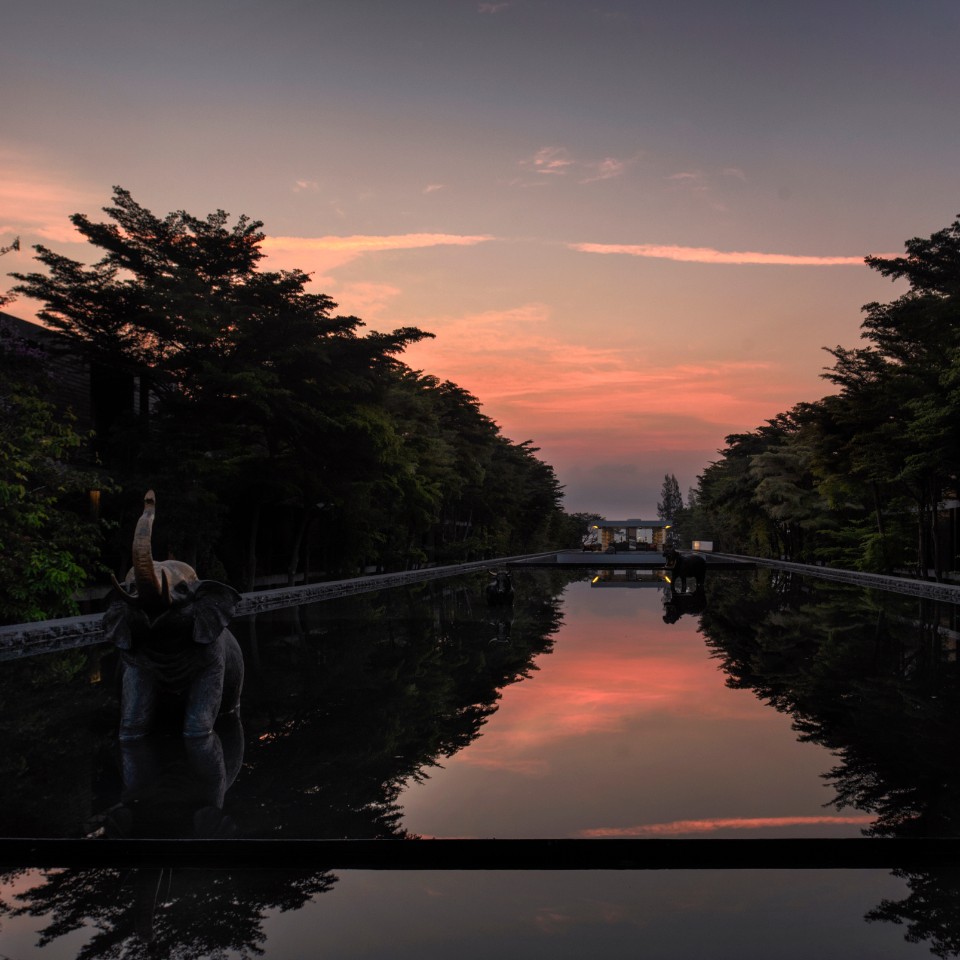 A tranquil pool reflects a vibrant pink and orange sunset, framed by dark trees. Elephant statues emerge from the water, adding a serene tone.