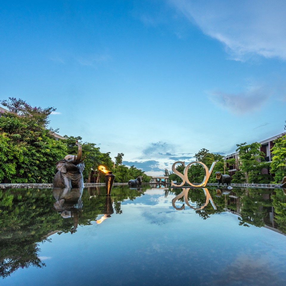 A serene water feature reflecting a clear blue sky, flanked by lush greenery. Elephant sculptures and a lit torch add elegance to the tranquil scene.